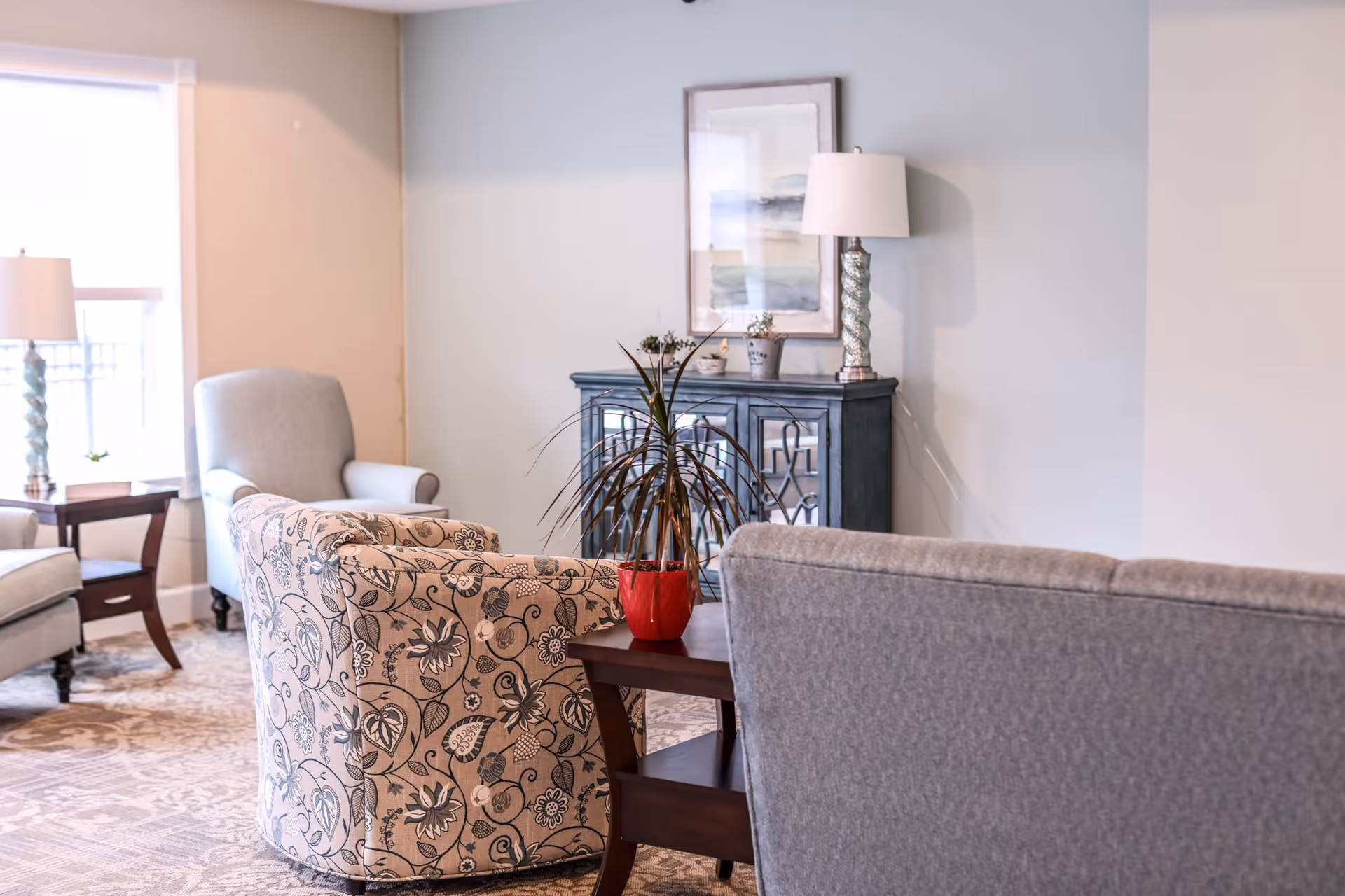 A cozy living room area with a patterned armchair, a gray sofa, and a light gray armchair near a window. There is a wooden side table with a red potted plant, a blue cabinet with decorative items and a lamp on top, and a framed abstract painting on the wall.
