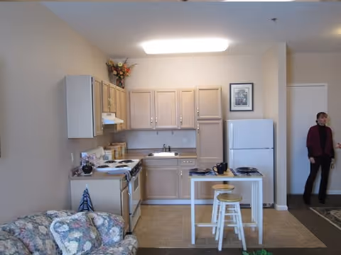 A small kitchen area in a senior living facility with light wood cabinets, a white refrigerator, stove, and sink. There is a small white table with two stools in front of the kitchen. A floral-patterned couch is partially visible on the left side. Two people are standing near a doorway on the right side of the image.