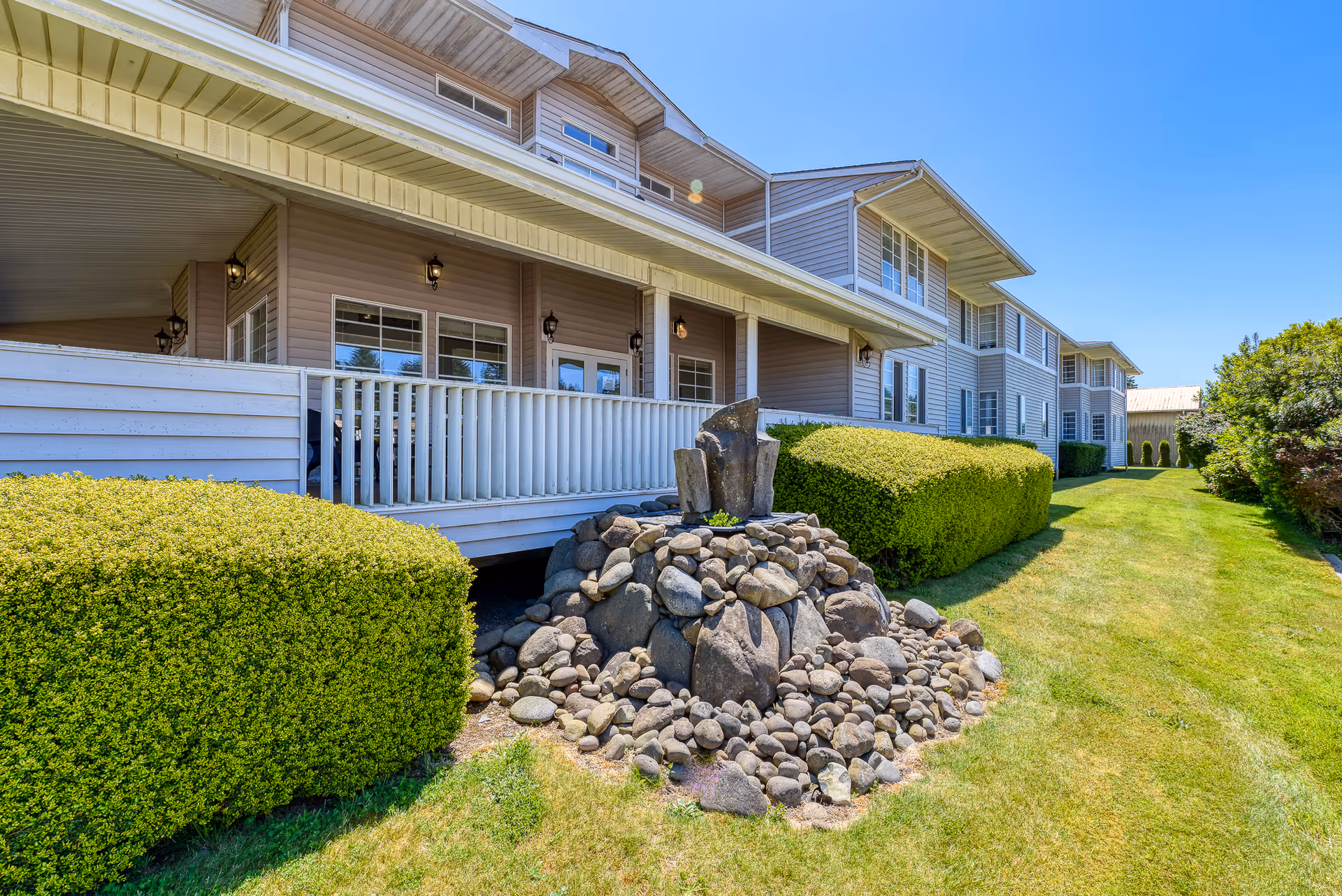 Exterior view of a multi-story assisted living facility with beige siding, white railings, and well-maintained landscaping including trimmed bushes and a rock water feature under a clear blue sky.