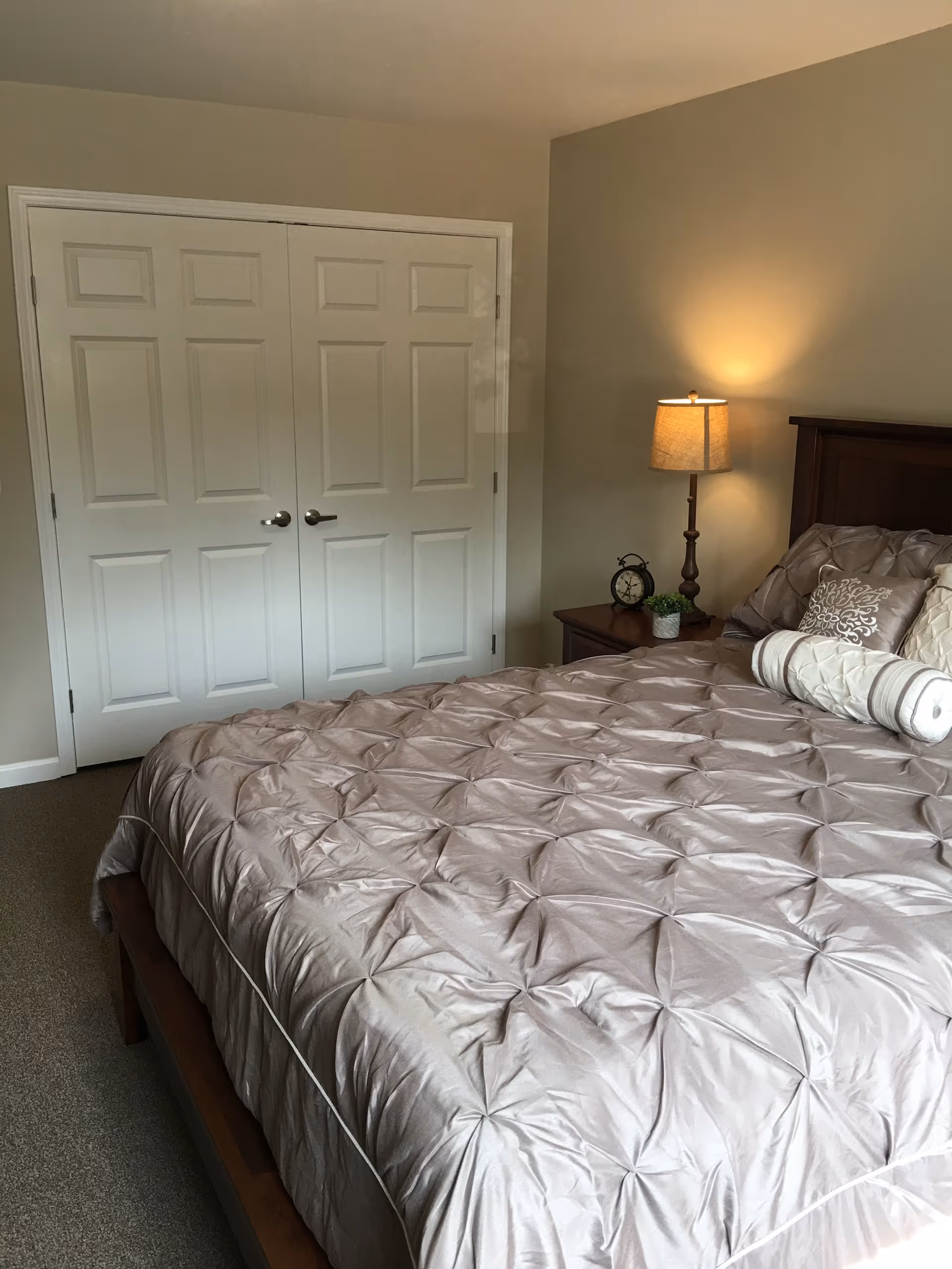 A bedroom with a large bed covered in a beige quilted comforter and several decorative pillows. Next to the bed is a wooden nightstand with a small lamp, an alarm clock, and a small potted plant. Behind the bed is a beige wall, and opposite the bed are white double doors with silver handles.