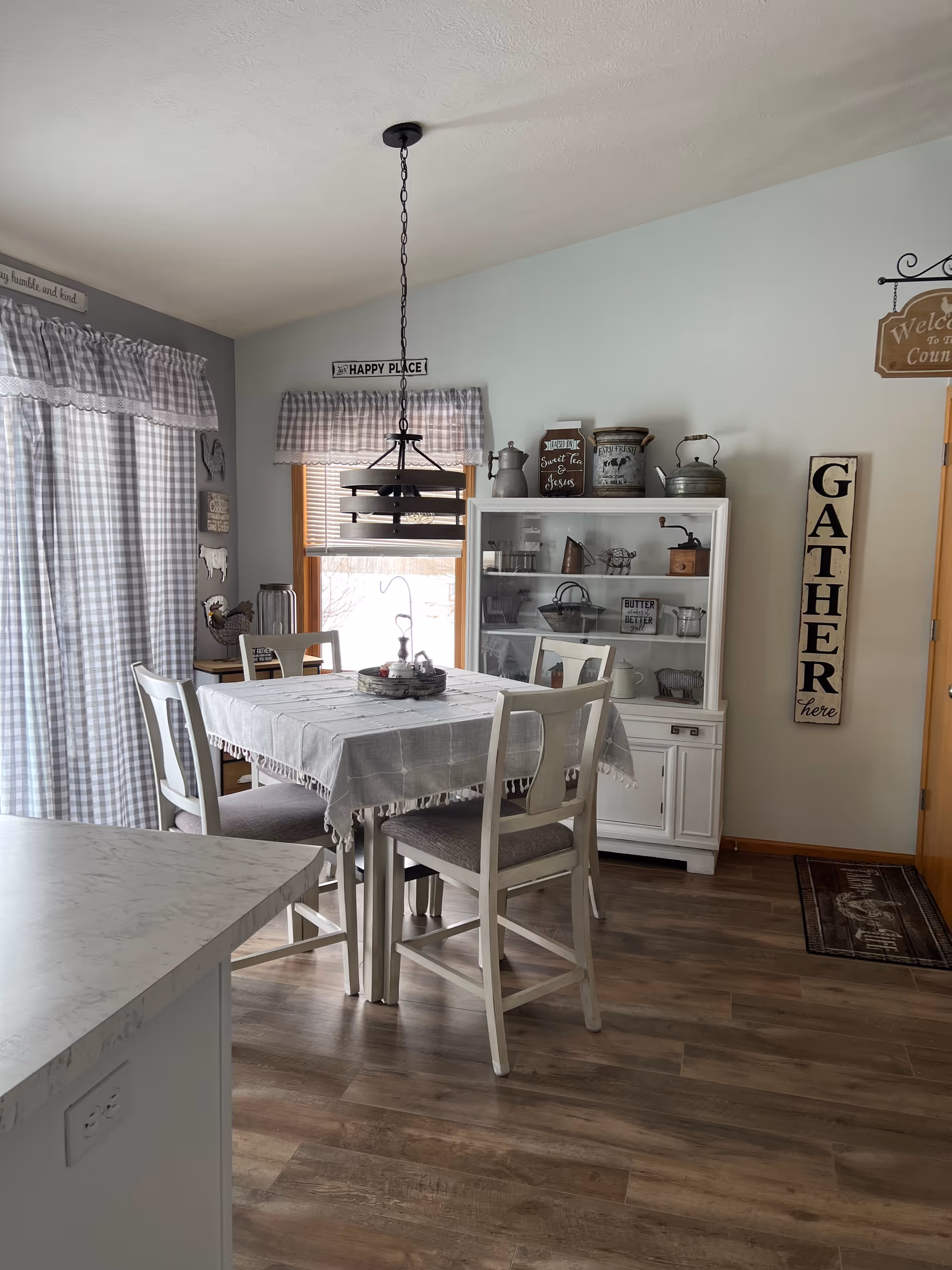 A cozy dining area with a square table covered by a gray tablecloth and surrounded by four white chairs with gray cushions. A black hanging light fixture is above the table. Behind the table is a white cabinet displaying various kitchen items and decor. The walls are light-colored with decorative signs, including one that says 'GATHER here' and another that says 'HAPPY PLACE'. A window with checkered curtains lets in natural light, and the floor is wood.