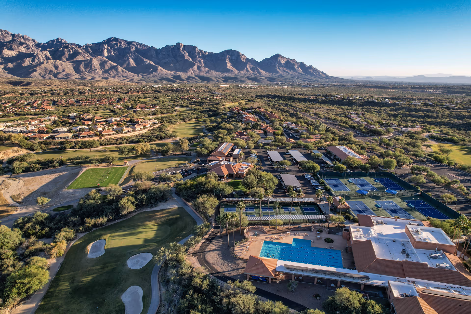Aerial view of Sun City Oro Valley Welcome Center showing a large swimming pool, tennis courts, parking areas, and surrounding desert landscape with mountains in the background under a clear blue sky.