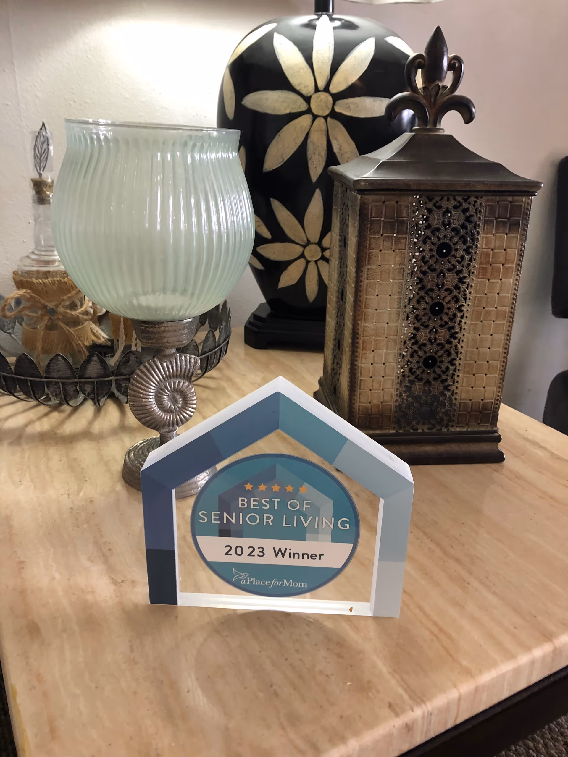 A decorative award plaque on a wooden table that reads 'Best of Senior Living 2023 Winner aPlaceforMom' surrounded by decorative items including a green glass candle holder, a black vase with white flower designs, and a brown ornate lantern.