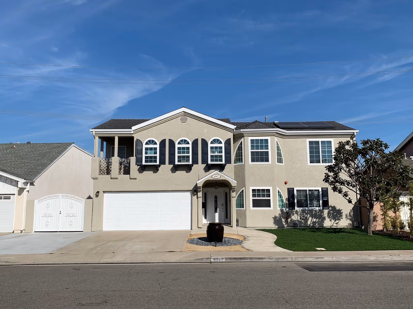 Front exterior view of a two-story beige residential building with a white garage door, arched windows with black shutters, a small balcony, a front entrance with a covered porch, a tree on the right side, and a clear blue sky above.