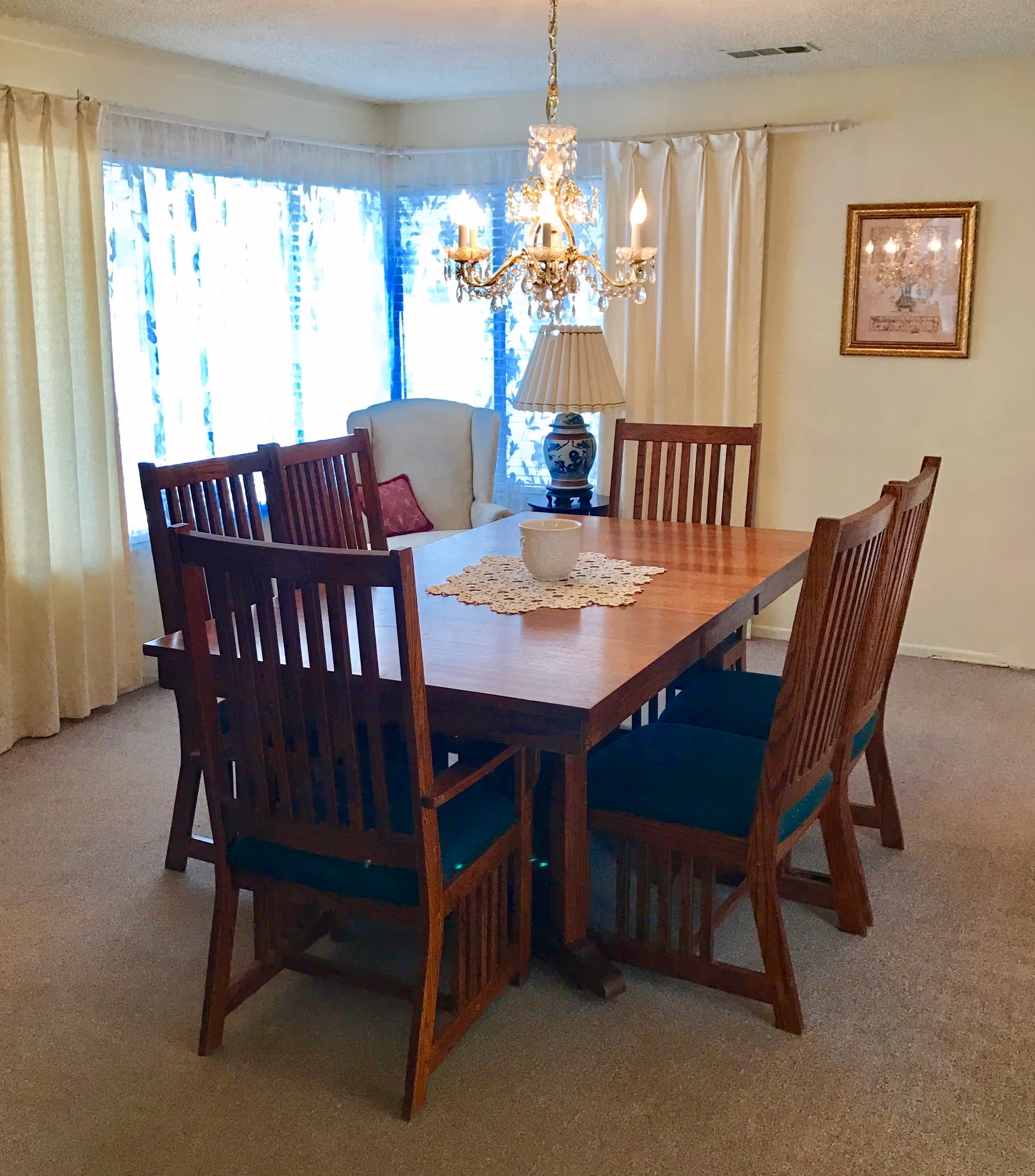 A dining room with a wooden dining table surrounded by six wooden chairs with green cushions. A white crocheted doily and a white cup are placed on the table. In the background, there is a white armchair with a red pillow, a table lamp with a blue and white base, cream-colored curtains, and a framed picture on the wall. A chandelier with lit bulbs hangs above the table.