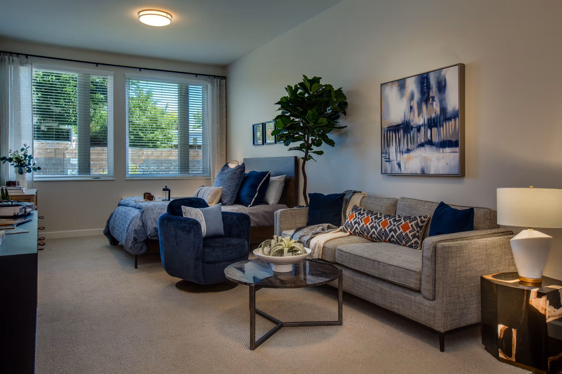A bright studio-style living area with a gray sofa, blue armchair, coffee table, and a bed near large windows.