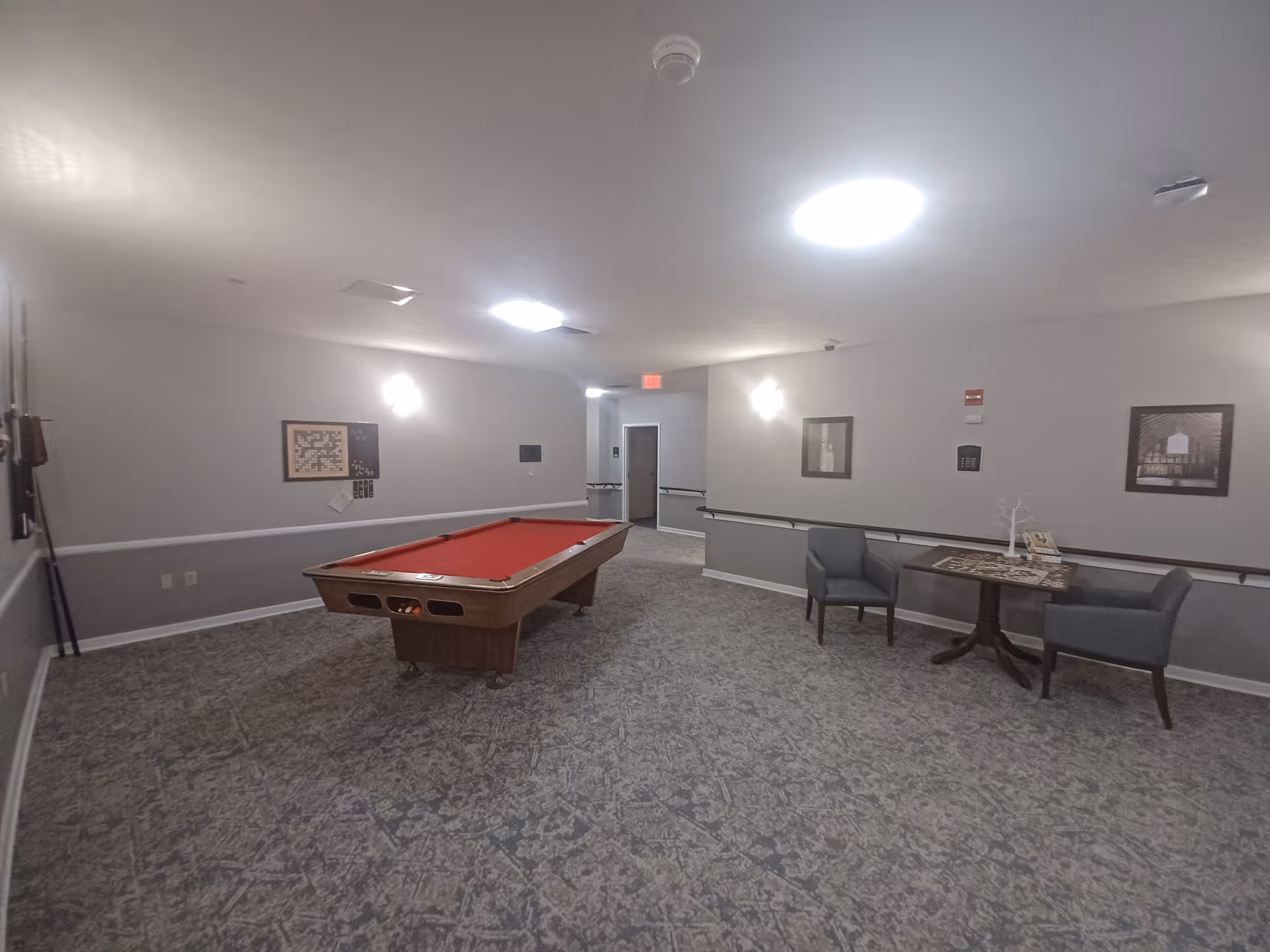 A recreational room with a red felt pool table in the center. The room has patterned carpet flooring, light gray walls with white trim, and ceiling lights. On the right side, there is a small table with two gray chairs and a board game on the table. The walls are decorated with framed pictures and a wall-mounted game board.