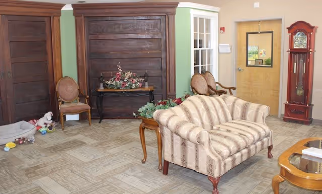 A cozy common room with an upholstered sofa, wooden chairs, decorative tables, and a grandfather clock.