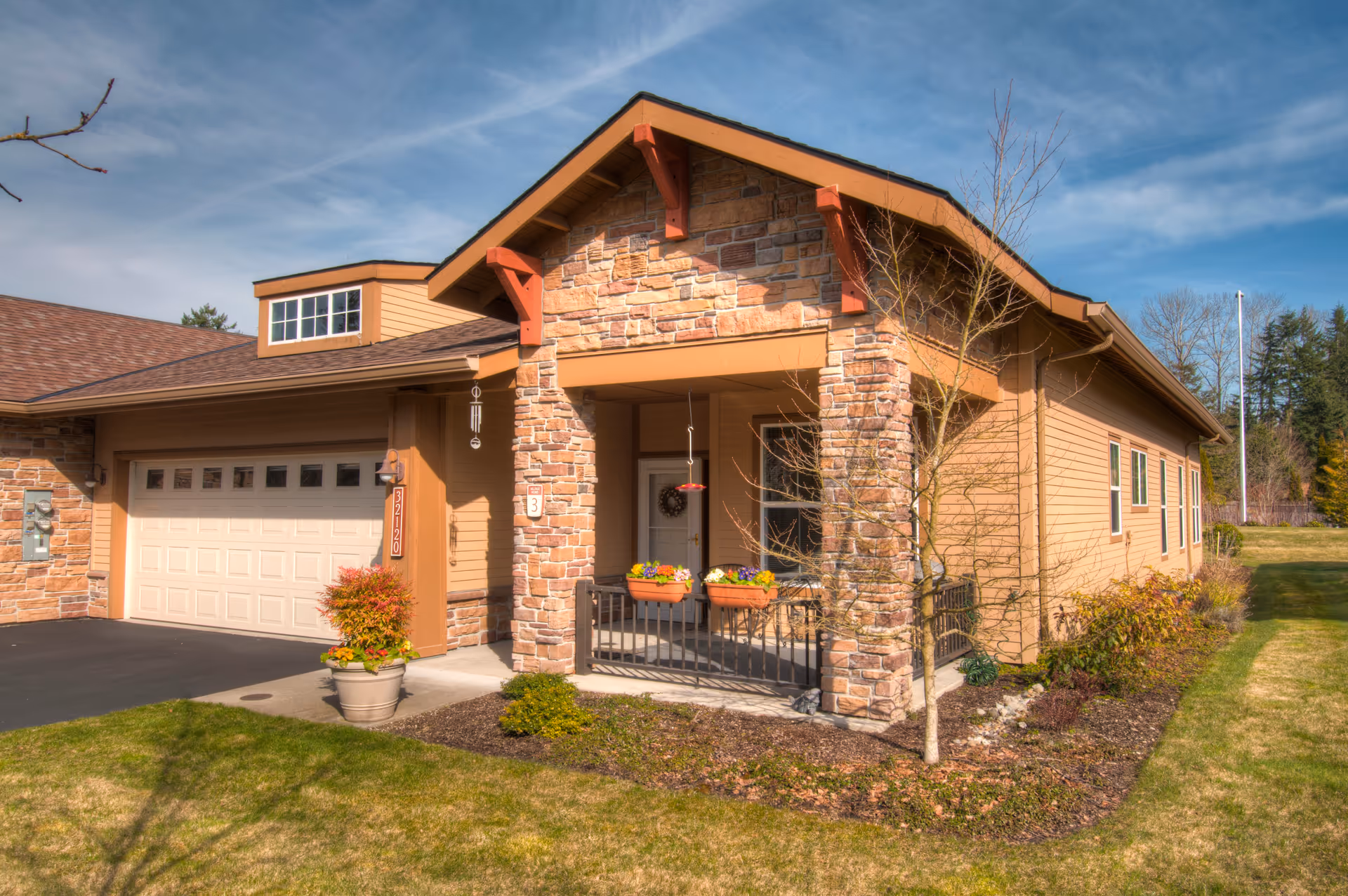 Exterior view of a single-story residential building with a stone and wood facade, a covered porch with flower pots, a garage, and a well-maintained lawn under a partly cloudy sky.