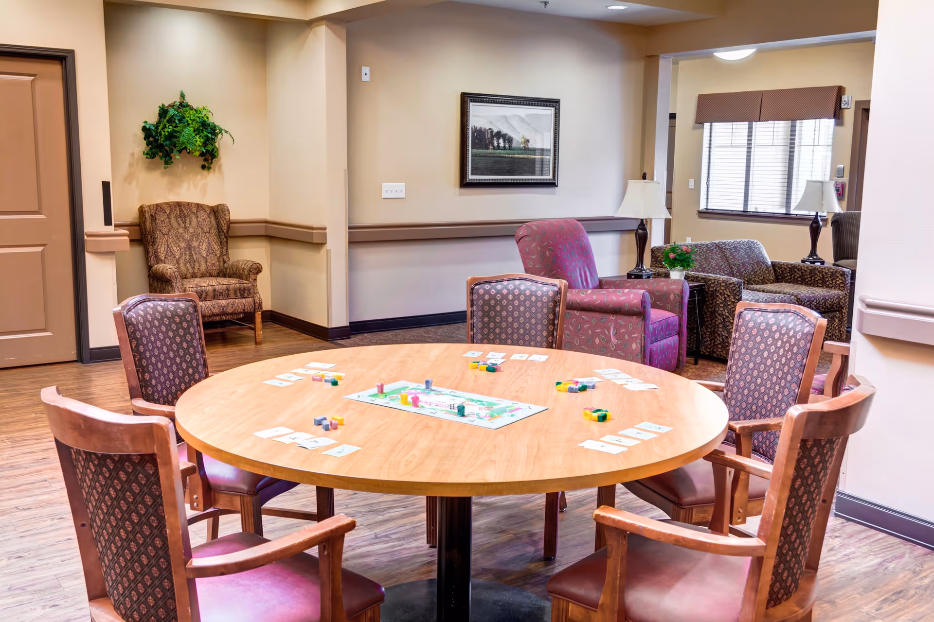 A cozy common area in a memory care residence featuring a round wooden table with six upholstered chairs arranged around it. On the table, there is a board game set up with cards and colorful game pieces. In the background, there are several armchairs, a framed picture on the wall, a potted plant, and a window with blinds allowing natural light to enter the room.