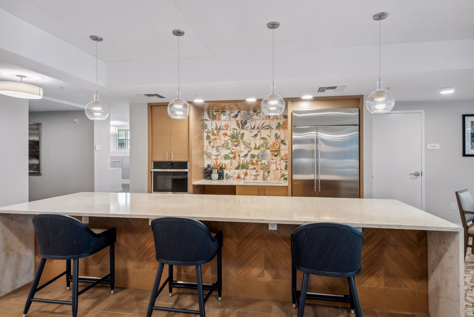 Communal kitchen with a marble island, three bar stools, pendant lights, stainless steel refrigerator, and a patterned tile backsplash.