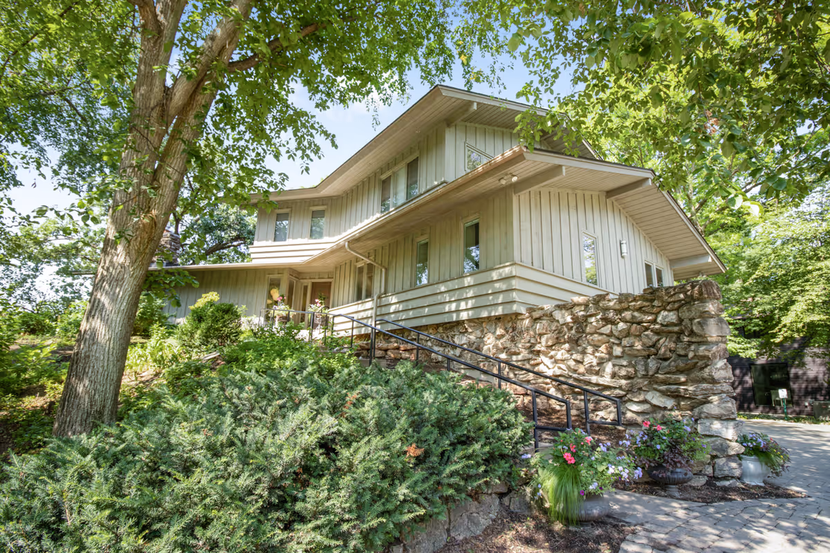 Exterior view of a two-story building with beige siding and a stone foundation, surrounded by lush green trees and shrubs. There is a paved walkway with a black handrail leading up to the entrance, and flower pots with colorful flowers are placed near the walkway.