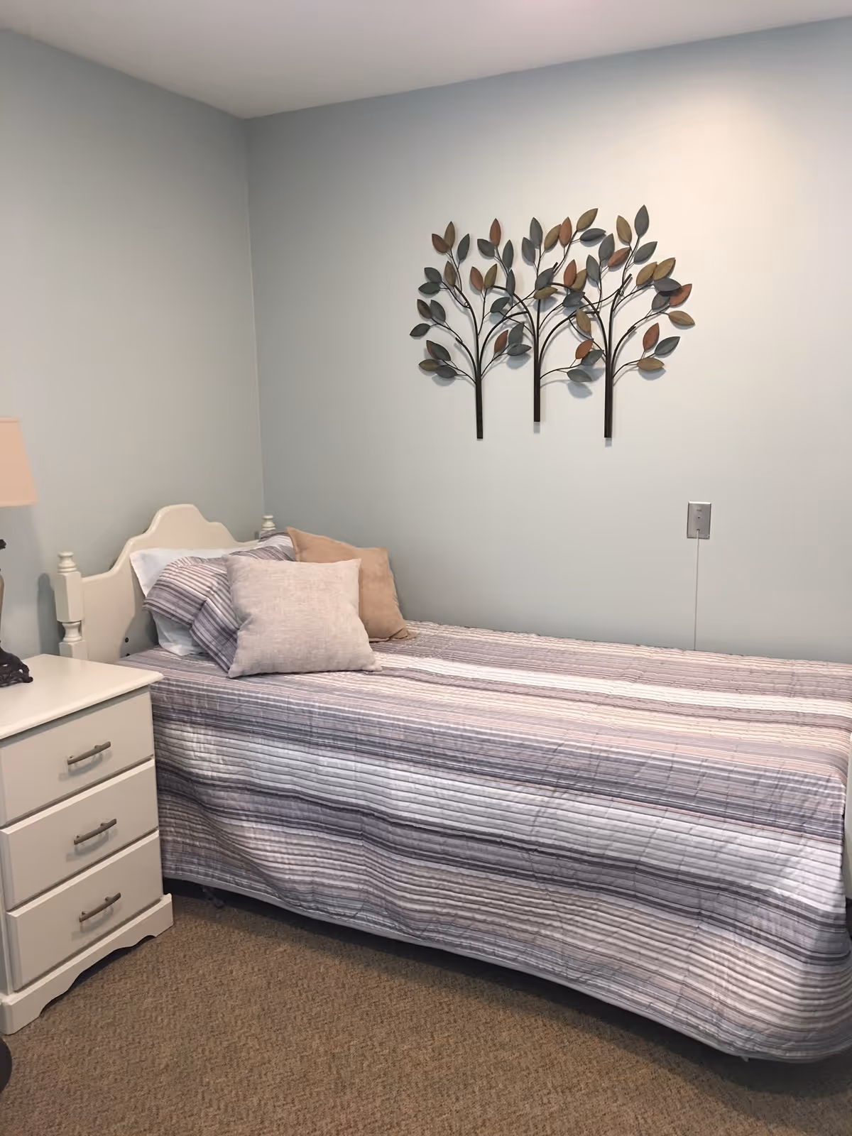 A small bedroom with a single bed covered in a striped bedspread and three pillows. Next to the bed is a white nightstand with three drawers and a lamp. On the wall above the bed is a decorative metal wall art piece featuring three stylized trees with leaves.