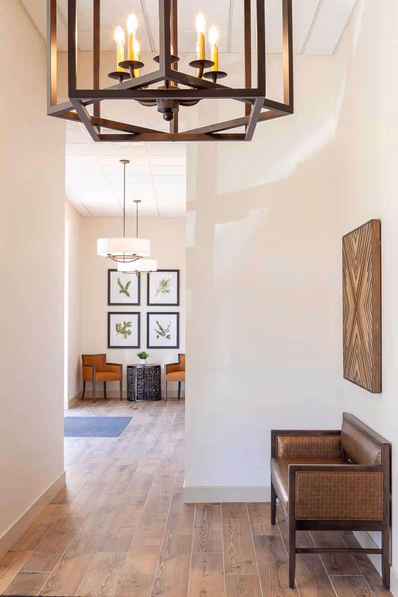 A bright interior hallway with wood floors, a pendant chandelier, a small seating area with framed botanical prints and chairs, and a bench against the wall.