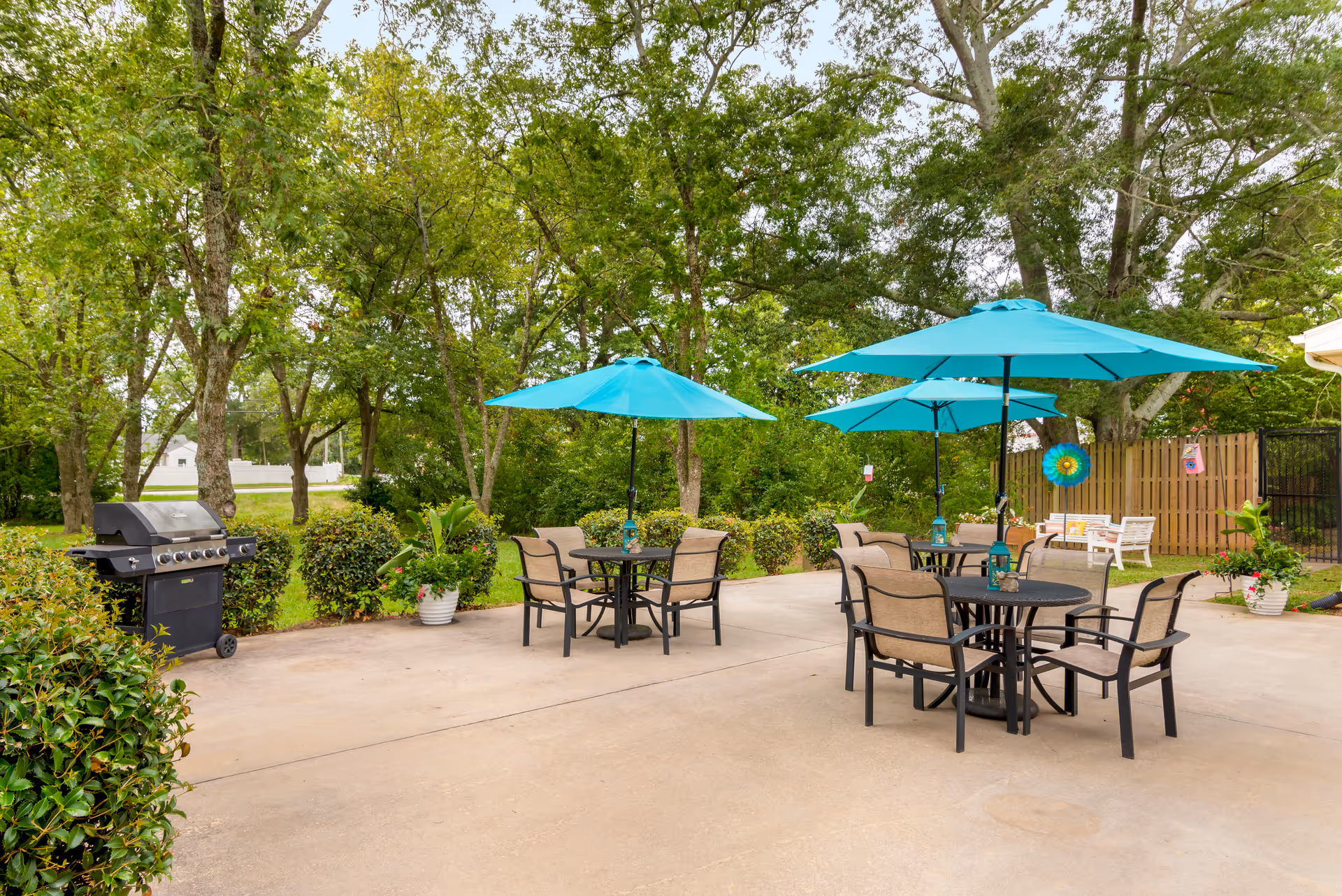 Outdoor patio area with several round tables and chairs under blue umbrellas, surrounded by greenery and trees. There is a black grill on the left side and some potted plants around the patio.