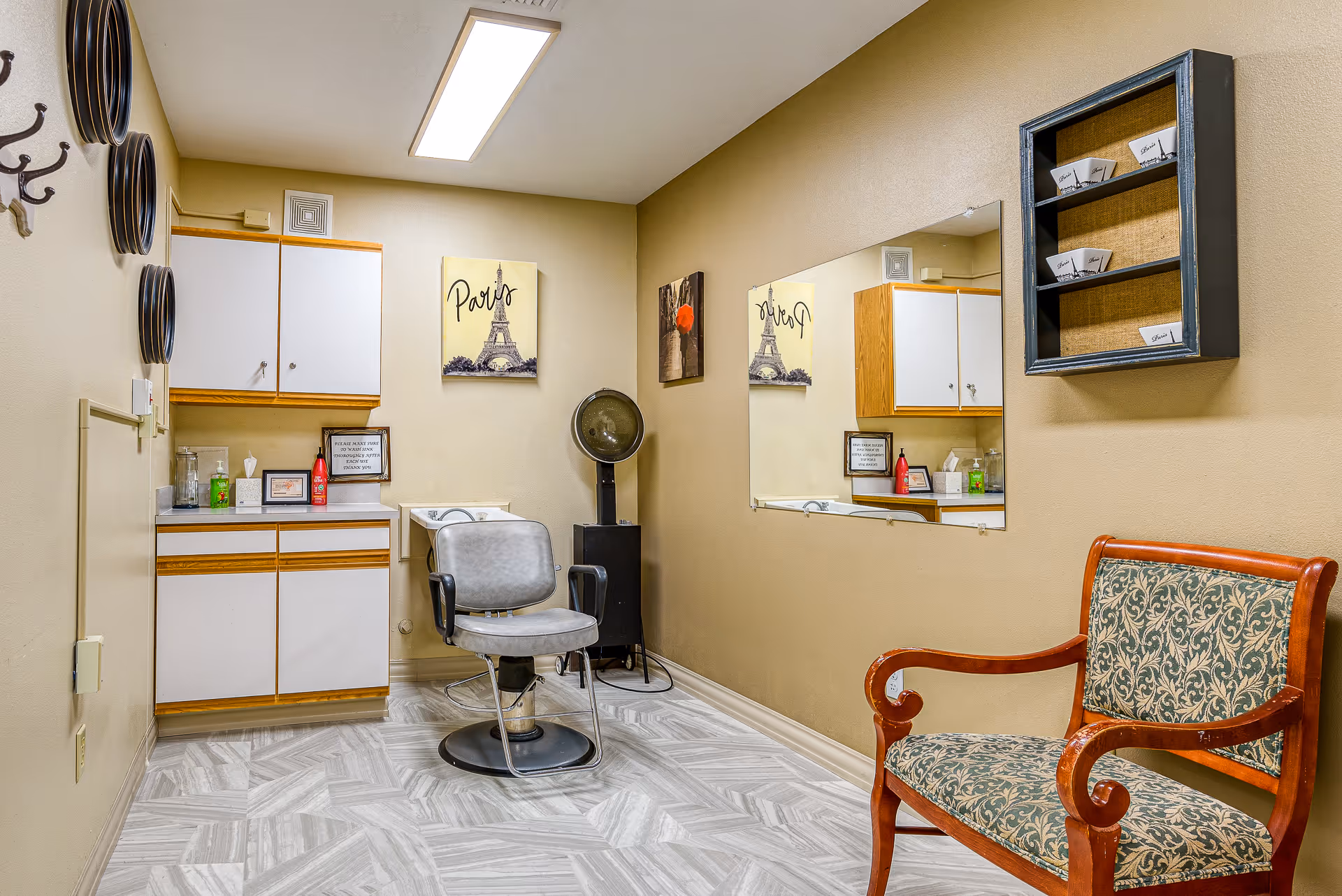 Interior of a small salon room in a senior living facility with a salon chair in front of a hair washing sink, a hair dryer, a wooden bench with patterned upholstery, wall-mounted cabinets, a large mirror, and decorative wall art including a Paris-themed picture.