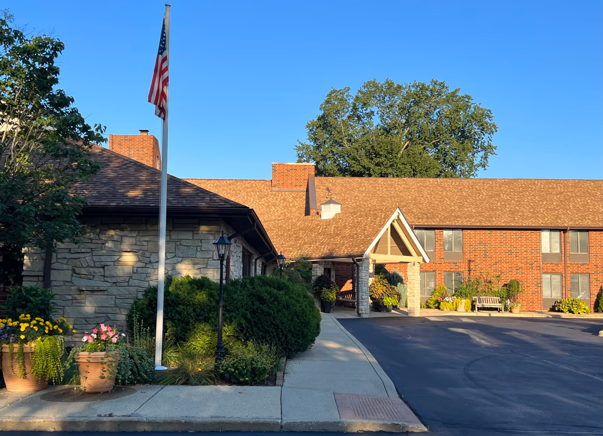 Front entrance of Independence Village showing a brick and stone building with a covered porte-cochère, American flag, and landscaped planters.