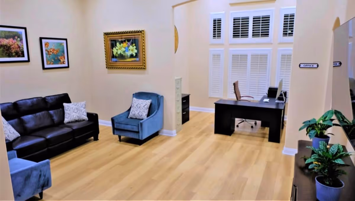 A bright office area in a care home with light wood flooring, beige walls, and white window shutters. The room features a black leather sofa, two blue armchairs with patterned cushions, framed floral paintings on the wall, a black desk with a computer and office chair, and several potted plants. A sign on the wall reads 'OFFICE'.