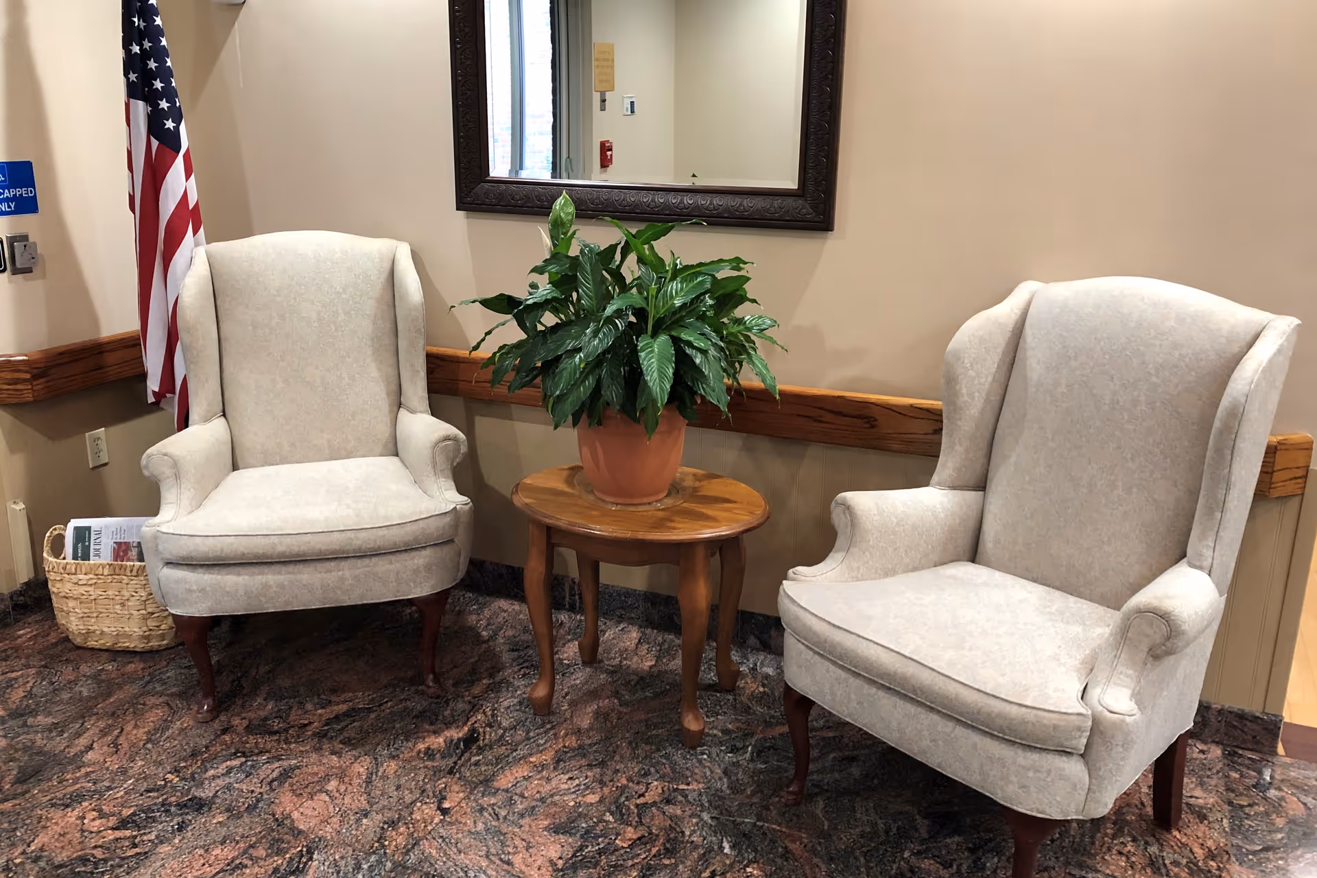 Two beige upholstered armchairs with wooden legs are positioned on either side of a small wooden table holding a potted green plant. Behind the table is a large framed mirror on a beige wall. An American flag stands in the corner next to a basket with magazines, and a blue handicapped sign is visible on the wall.