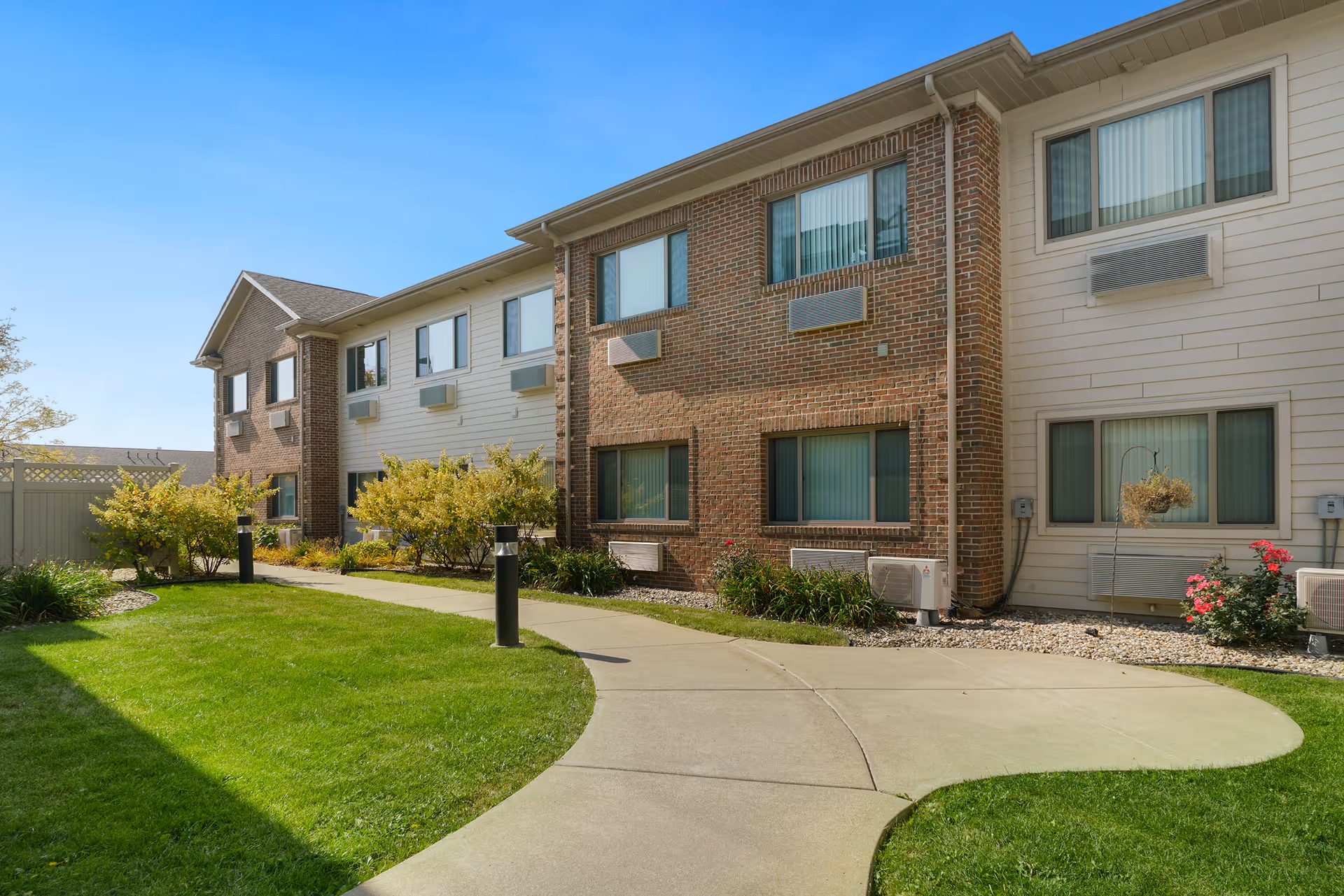 Exterior view of a two-story senior living facility building with brick and siding walls, multiple windows with air conditioning units, a curved concrete walkway, green grass, and landscaped bushes under a clear blue sky.