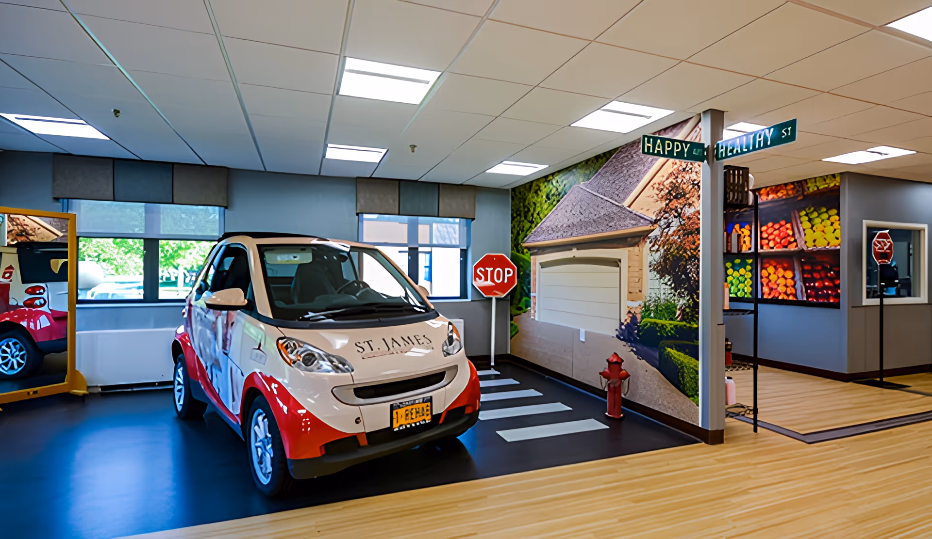 Indoor room designed to look like a small street scene with a small car labeled 'St. James' parked on a black floor with white crosswalk lines. There is a stop sign, a fire hydrant, and street signs reading 'Happy' and 'Healthy St'. The walls have large murals depicting a house and colorful produce. A large mirror reflects part of the scene.