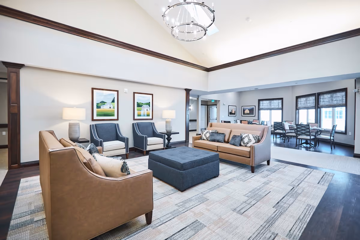 A spacious and well-lit living room area in Cedarhurst of Lebanon featuring two brown leather sofas with decorative pillows, two blue armchairs, a large blue ottoman in the center, and two table lamps on side tables. The room has high ceilings with a modern chandelier, framed landscape paintings on the walls, and large windows with shades. In the background, there is a dining area with multiple tables and chairs.