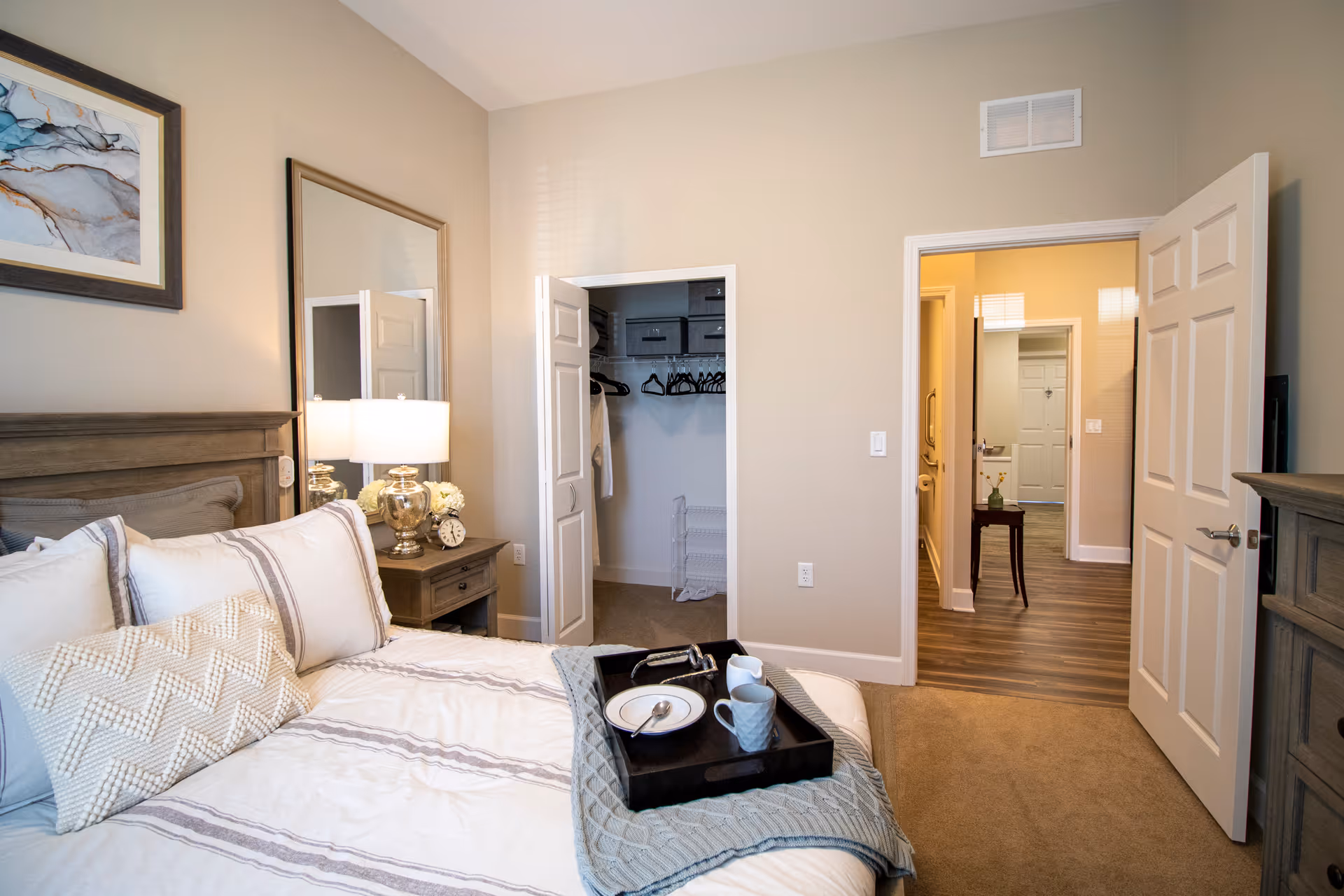Furnished bedroom with a made bed, nightstand and lamp, an open closet, and a view into a hallway.