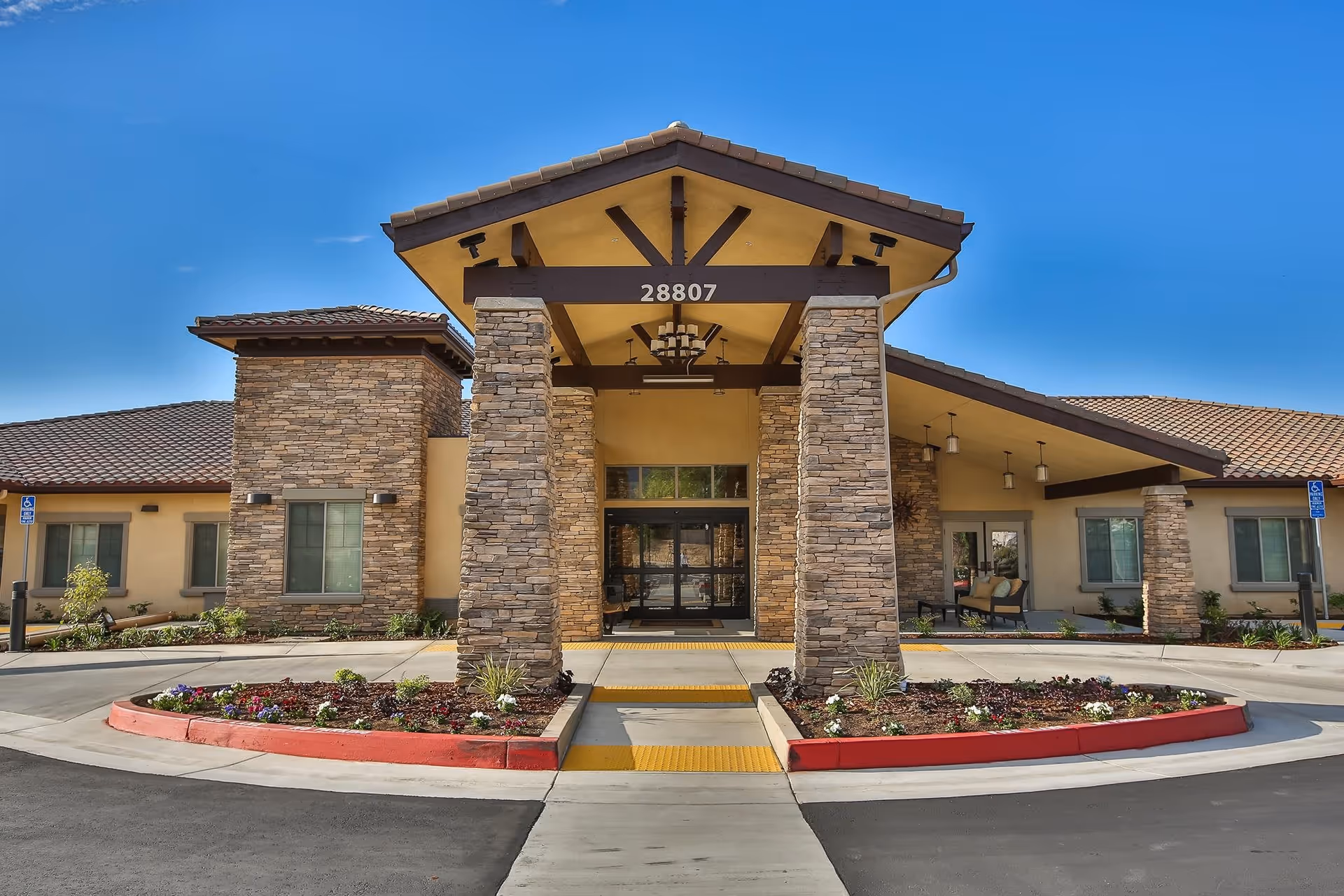 Front exterior view of a senior living facility with stone pillars supporting a covered entrance. The building has a tiled roof and beige walls with stone accents. There are flower beds with colorful flowers in front of the entrance and a clear blue sky above.