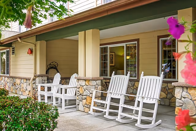 Covered outdoor patio area at Brookdale Arbor Place with white wooden rocking chairs arranged along a stone and wood exterior wall, surrounded by green bushes and pink flowers.