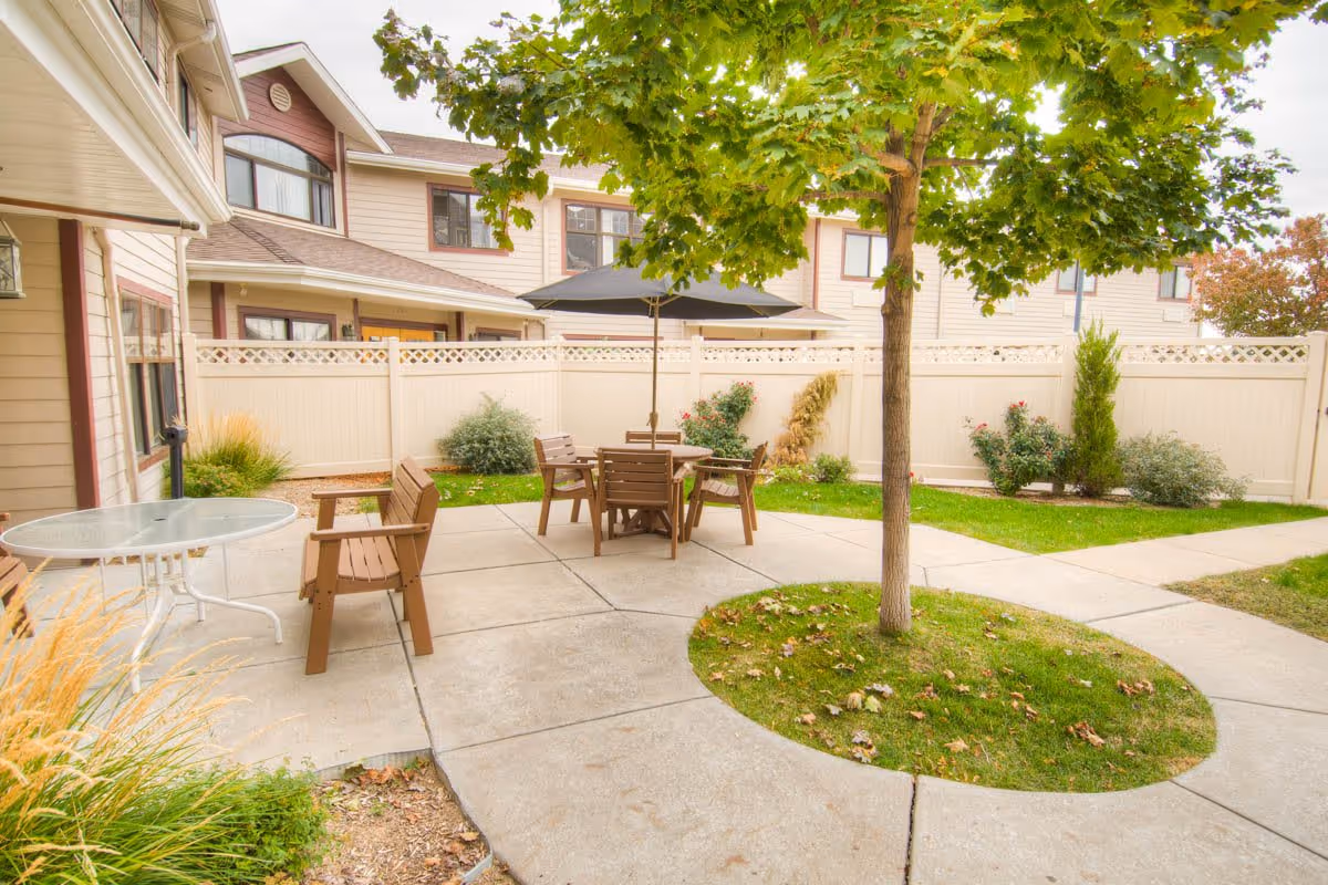 Outdoor patio area with a circular concrete walkway surrounding a tree, wooden chairs and a table with an umbrella, adjacent to a beige building with white fencing and some greenery.