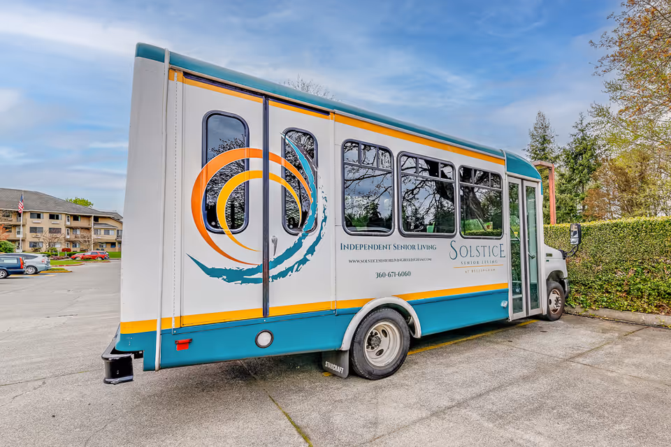 A teal and white shuttle bus parked in a parking lot near a senior living facility. The bus has the logo and branding for Solstice Senior Living at Bellingham, including text that reads 'Independent Senior Living' along with a phone number and website. In the background, there are residential buildings, cars, trees, and a partly cloudy sky.