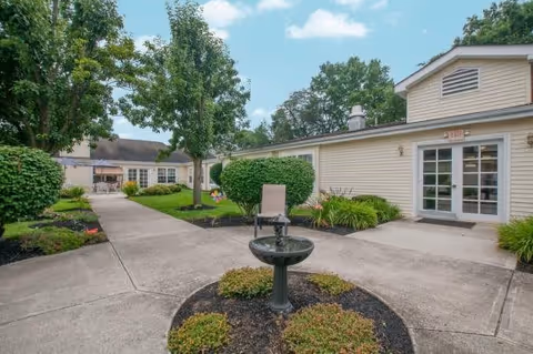 Outdoor courtyard area at The Courtyard at Newark featuring a concrete walkway, a small water fountain surrounded by shrubs, manicured bushes, trees, and beige buildings with white trim under a partly cloudy sky.