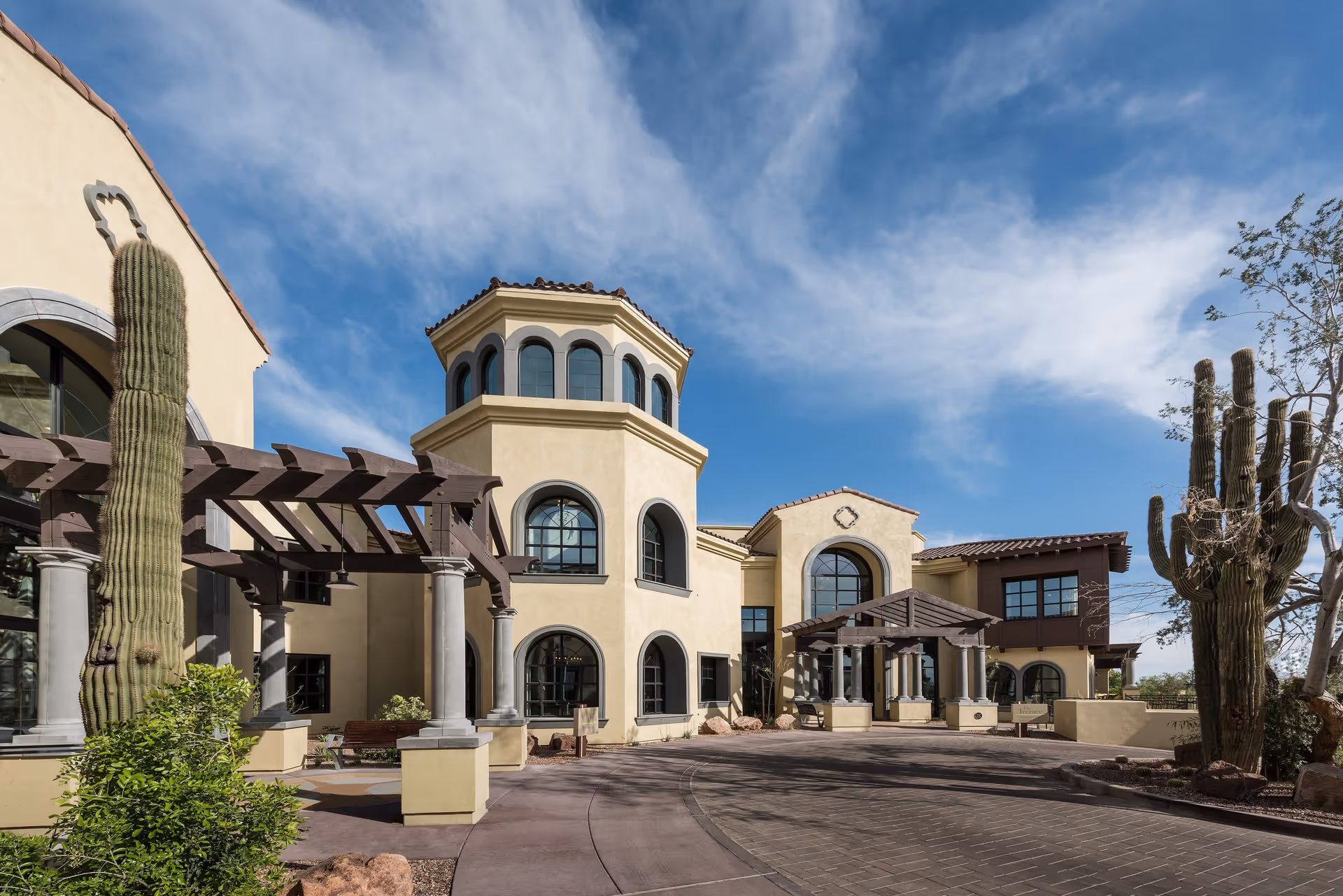 Exterior view of a Mediterranean-style building with beige stucco walls, arched windows, and a tower-like structure. The entrance features a covered walkway with wooden beams and stone columns. There are tall cacti and desert landscaping around the building under a partly cloudy blue sky.