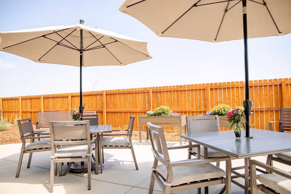Outdoor patio area with tables and chairs under large beige umbrellas, surrounded by a wooden fence and flower planters.