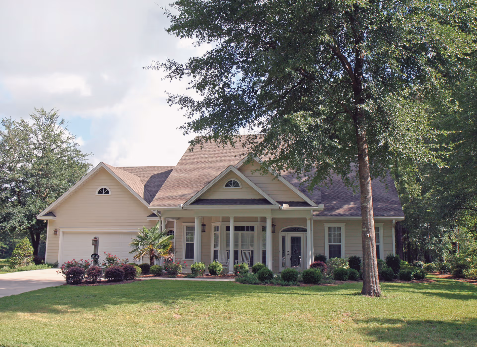 A single-story beige house with a front porch featuring two rocking chairs, surrounded by green grass, bushes, and trees under a partly cloudy sky.