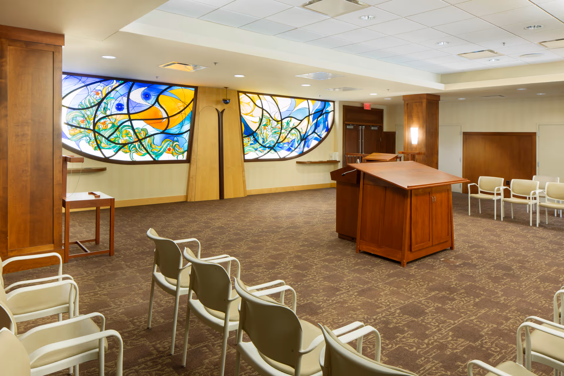 Spacious interior meeting/chapel room with stained-glass windows, rows of chairs, and a wooden lectern.