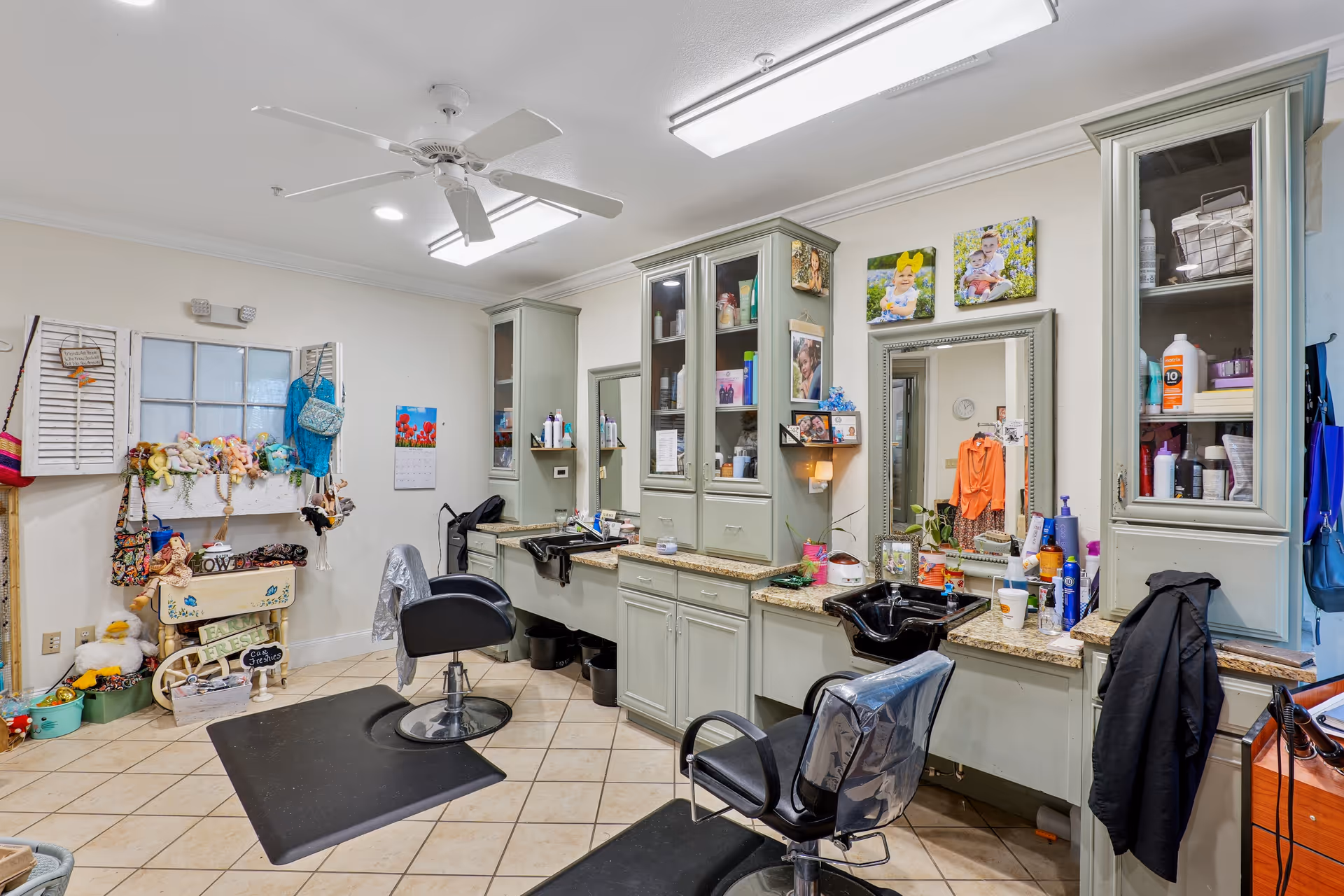 Interior of a salon area with two black salon chairs in front of a counter with sinks and mirrors. The counter has various hair and beauty products. Cabinets with glass doors hold additional supplies. On the left side, there is a wall with hanging bags, stuffed animals, and decorative items. The floor is tiled and the ceiling has a white fan and recessed lighting.