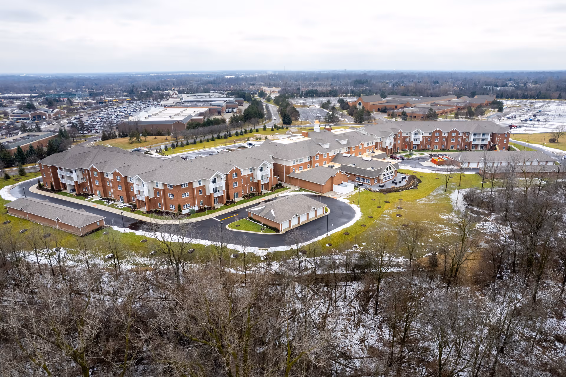 Aerial view of a large brick retirement community complex with multiple connected buildings, driveways, parking areas, lawns with patches of snow, and surrounding trees.