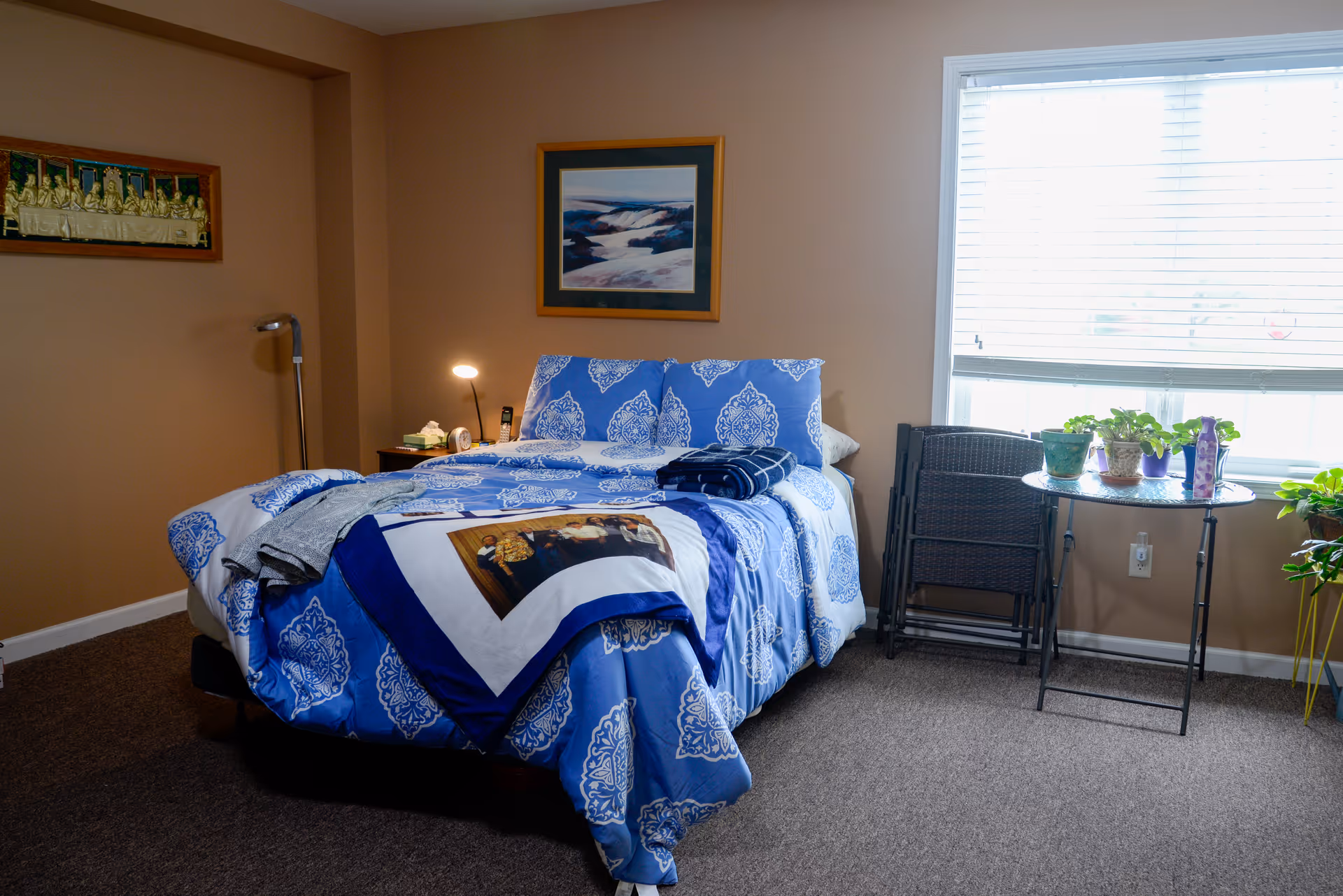 A bedroom featuring a bed with blue patterned bedding, a nightstand and lamp, framed artwork on the wall, and a window-side table with potted plants.