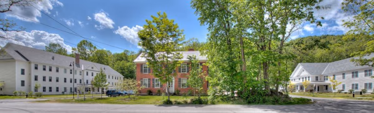 Panoramic view of West River Valley Assisted Living Residence showing multiple buildings surrounded by trees and greenery under a partly cloudy blue sky.