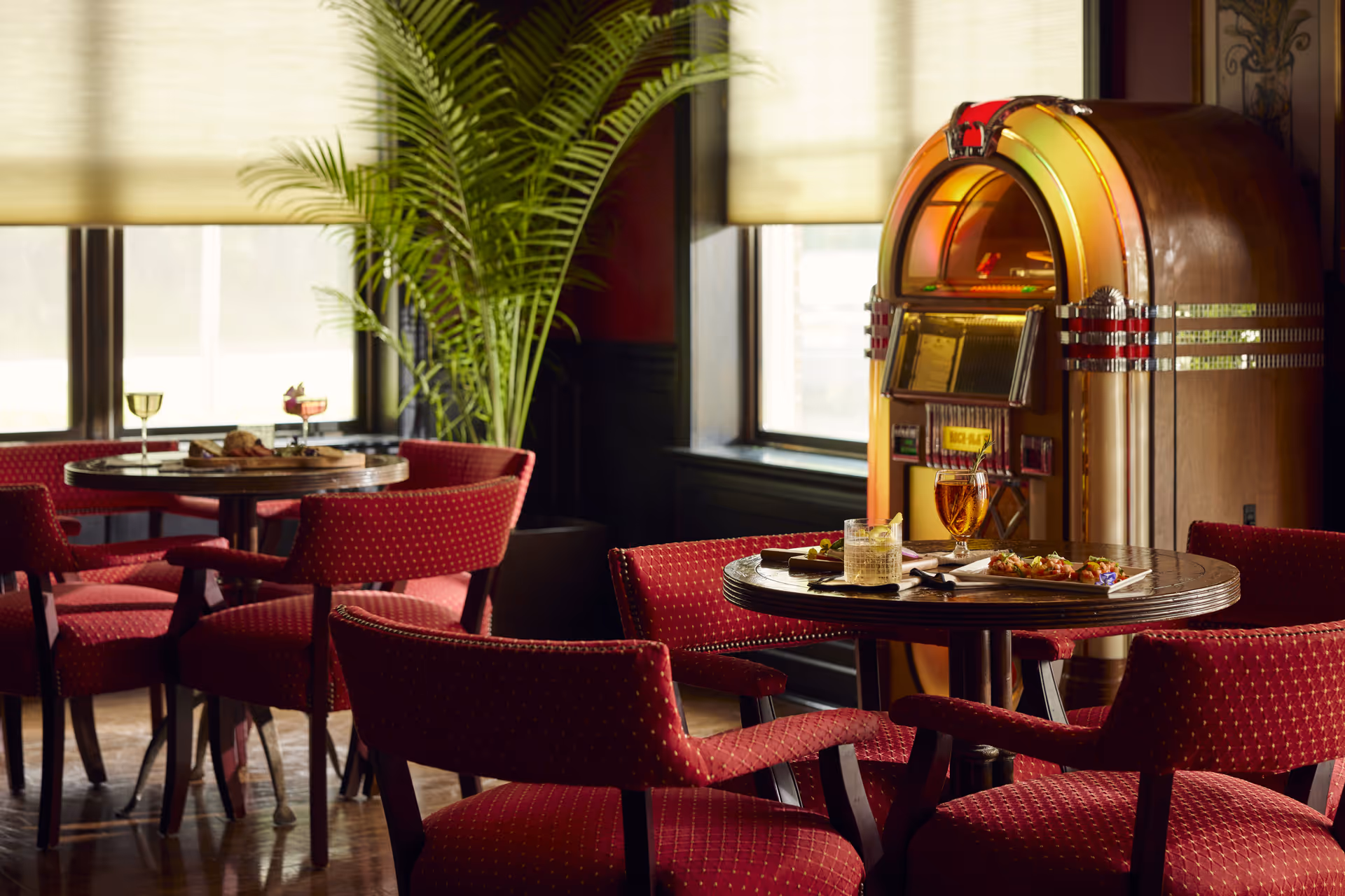 A cozy dining area with round wooden tables and red upholstered chairs with a diamond pattern. On one table, there are two drinks and a plate of appetizers. In the background, there is a vintage jukebox and a large green potted plant near windows with beige blinds.