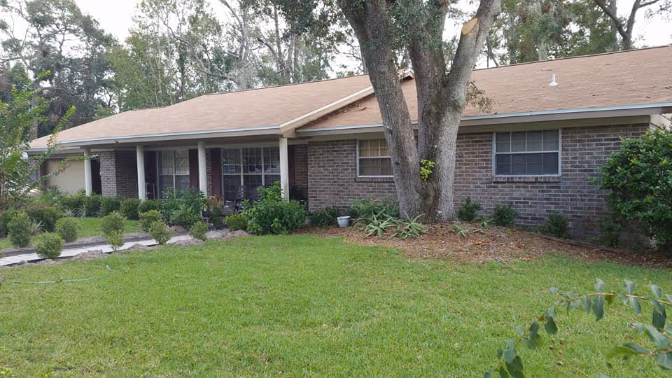 Single-story brick building with a brown shingled roof, surrounded by green grass, shrubs, and trees in the front yard under a clear sky.