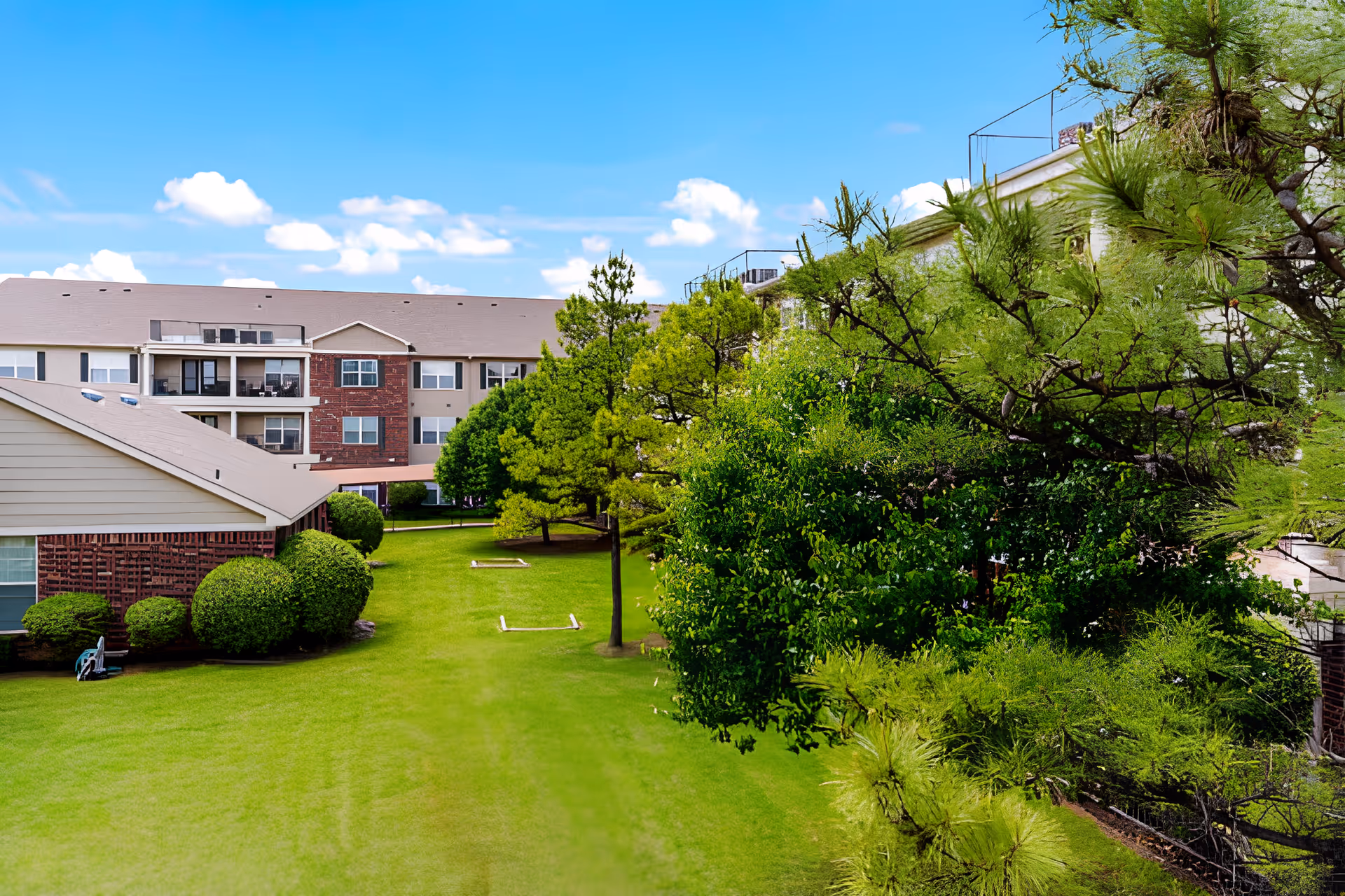 A well-maintained outdoor garden area at Elison Independent Living of Statesman Club featuring green grass, neatly trimmed bushes, and several trees with a multi-story residential building in the background under a blue sky with scattered clouds.