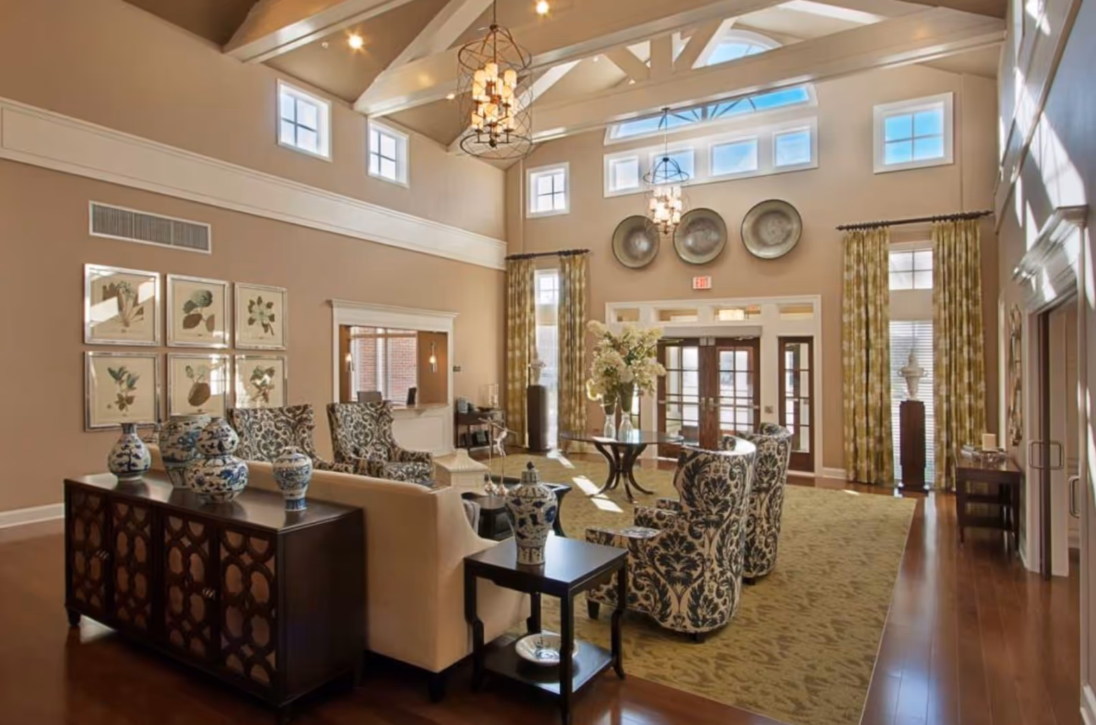 Spacious and well-lit living room area in Crestview Retirement Community featuring high vaulted ceilings with exposed beams, multiple windows allowing natural light, patterned armchairs, a beige sofa, decorative vases on dark wooden furniture, a glass-top table with a flower arrangement, and large curtains framing the entrance doors.