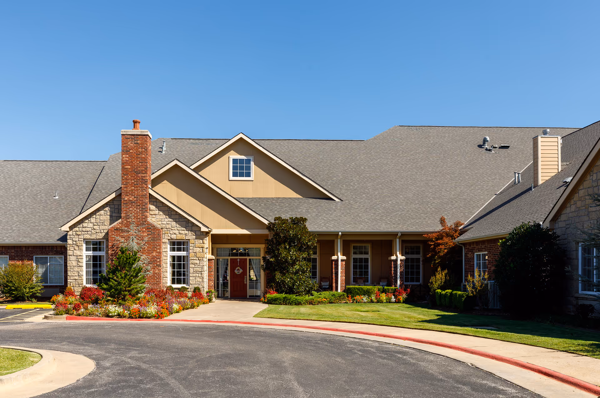 Front exterior view of a single-story assisted living facility building with a large chimney, multiple windows, a red front door, well-maintained landscaping with colorful flowers, shrubs, and a curved driveway under a clear blue sky.
