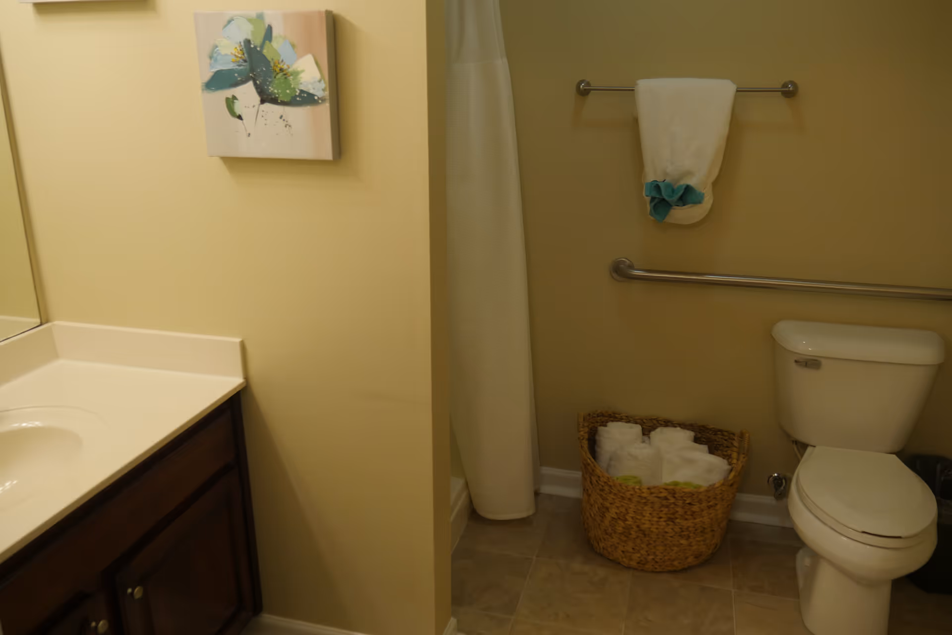 Bathroom with a sink and vanity on the left, a shower curtain in the center, and a toilet, towel rack, and wicker basket of towels on the right.