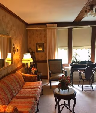A cozy living room with a patterned orange sofa, a wooden side table with a lamp, an armchair, a round wooden coffee table with a floral arrangement, and a desk with a chair near two windows with blinds partially drawn. The walls have a vintage floral wallpaper and there is a large mirror above the sofa.
