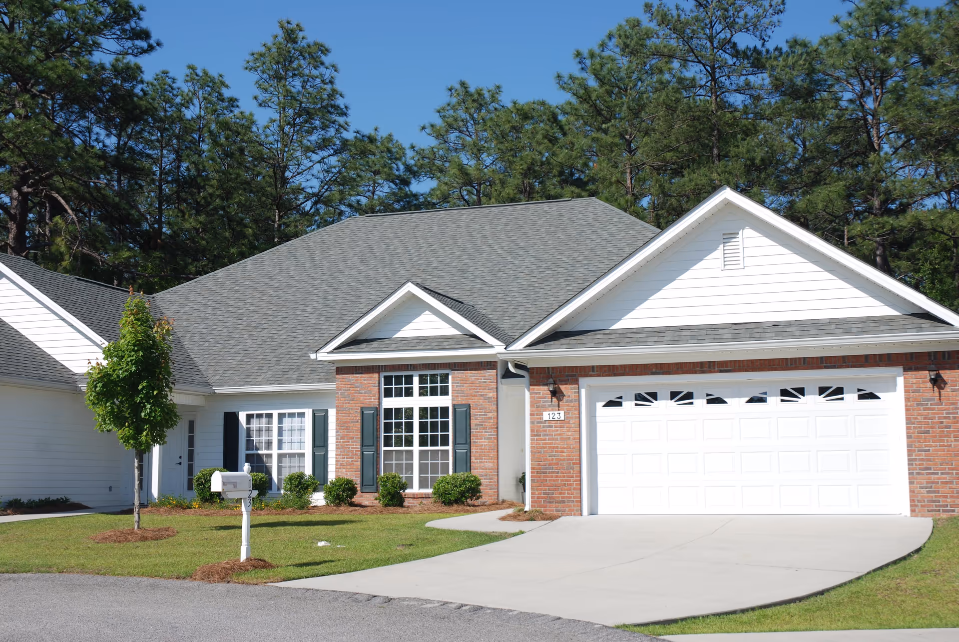 A single-story residential building with a gray shingled roof, white siding, and red brick accents. The house features a large white garage door, a front window with black shutters, a small tree, and a white mailbox with the number 123. The driveway and lawn are well-maintained, and tall pine trees are visible in the background under a clear blue sky.