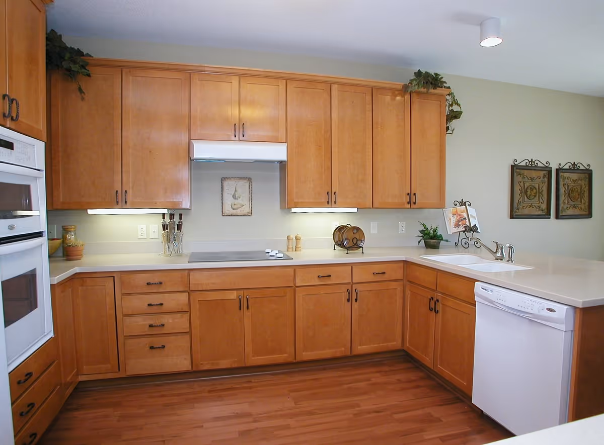 A modern kitchen with wooden cabinets, a white oven, a stovetop with a range hood, a white dishwasher, and a sink. The countertop is light-colored, and there are decorative items such as plants and framed pictures on the walls and countertop.