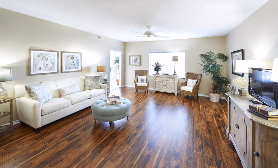 A bright and spacious living room with wooden flooring, a white sofa with decorative pillows, a round blue ottoman, two wicker chairs with cushions, a wooden cabinet with a TV on top, a large potted plant, framed artwork on the walls, and a ceiling fan.